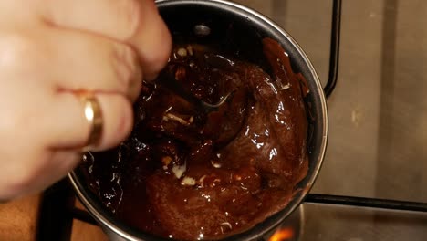 una barra de chocolate con leche rota con avellanas mezcladas con mantequilla se derrite en la estufa de gas mientras una mujer con anillo de matrimonio se agita en una olla pequeña para derretir