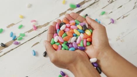 Video-of-overhead-view-of-biracial-man-holding-multi-coloured-sweets-over-white-rustic-background