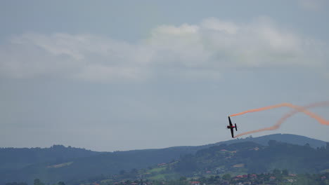 single airplane with orange smoke trail performing aerial stunt above green hills during airshow