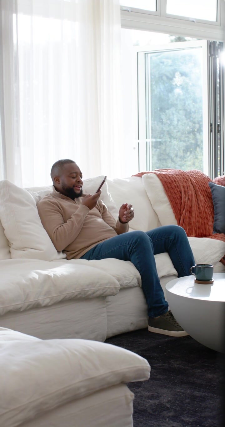 Video vertical: hombre adulto leyendo un mensaje en su teléfono inteligente, sonriendo y luego respondiendo una llamada en un sofá blanco