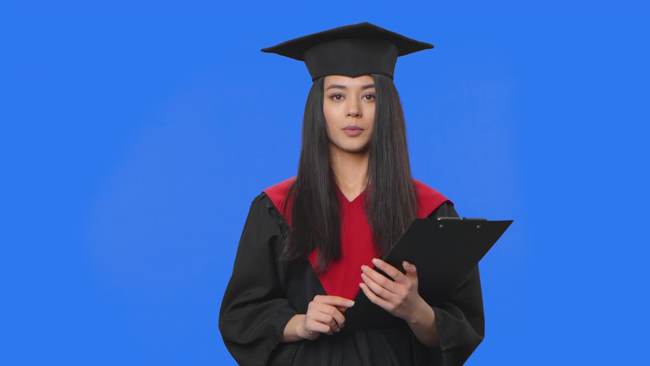 retrato de una estudiante en traje de graduación, contando información y sosteniendo una carpeta negra en las manos. mujer joven posando en el estudio con fondo de pantalla azul. primer plano. cámara lenta lista 59.94fps