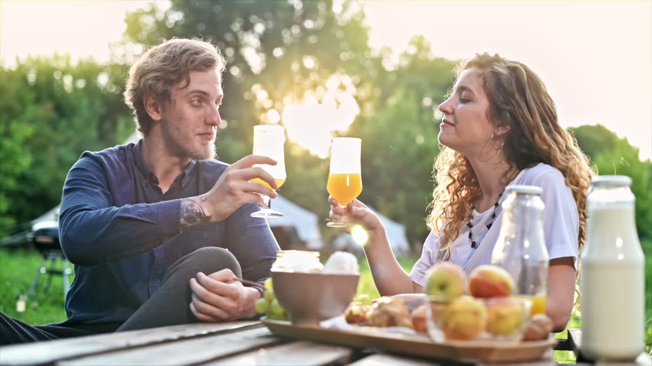 A couple having breakfast in the nature at glamping. Drinks, food, talking and smiling. Tent on the background. Slow motion