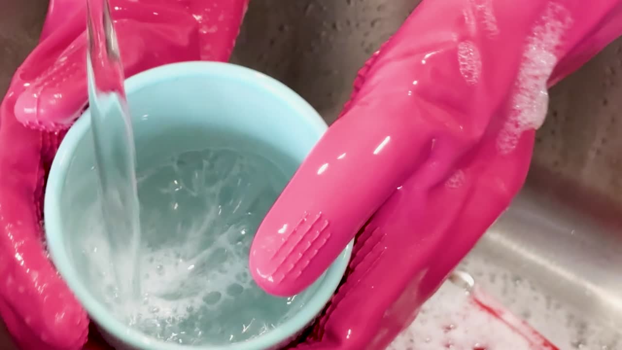 Mani che indossano guanti di gomma rosa strofinano e risciacquano una tazza di ceramica blu sotto l'acqua corrente