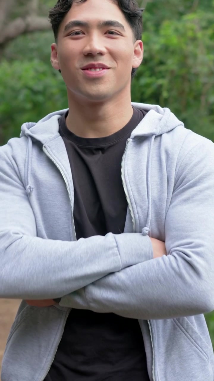 세로 영상: 카메라 줌에 반응하며 팔짱을 끼고 웃는 후드티를 입은 성인 아시아 남성이 오솔길에 있는 모습