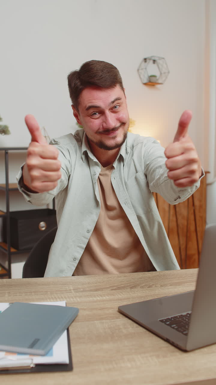 Uomo caucasico felice che guarda con approvazione la telecamera mostrando il pollice in su, seduto alla scrivania dell'ufficio di casa.