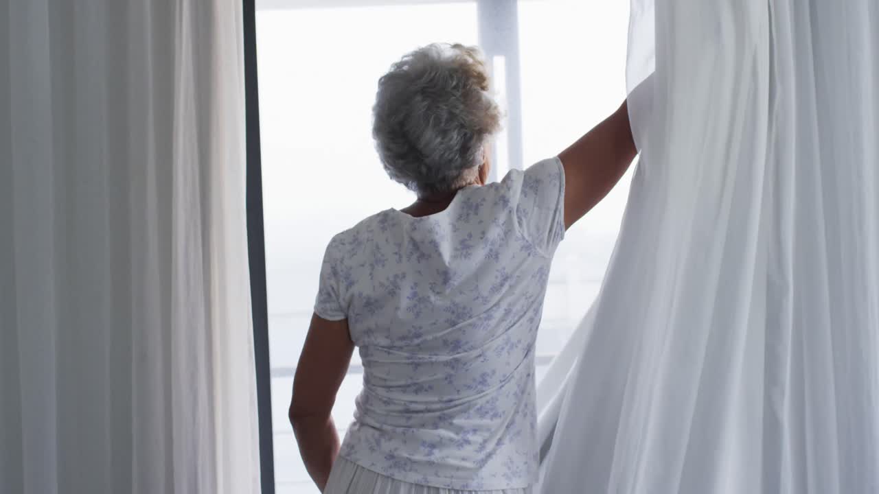 Anziana donna afroamericana che apre le tende della sua finestra a casa.
