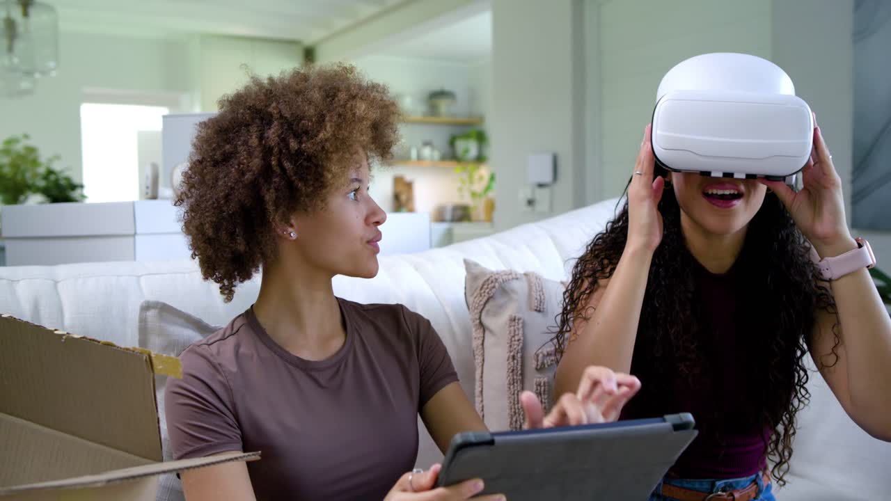 African American women unboxing VR headset leading right woman fitting it left using tablet on sofa
