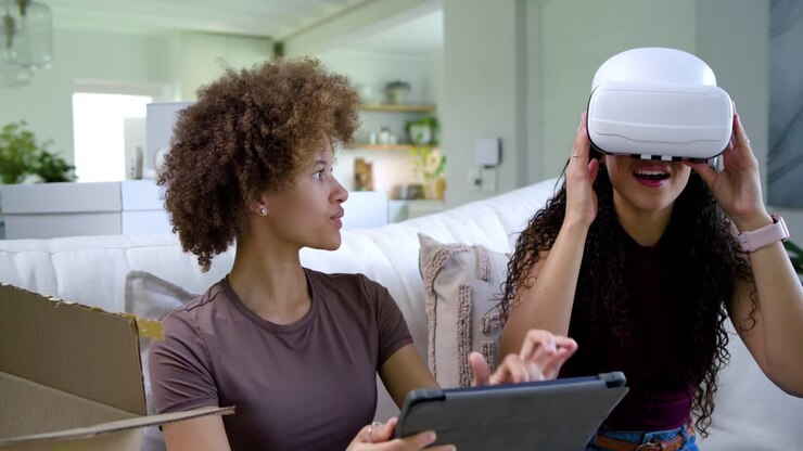African American women unboxing VR headset leading right woman fitting it left using tablet on sofa