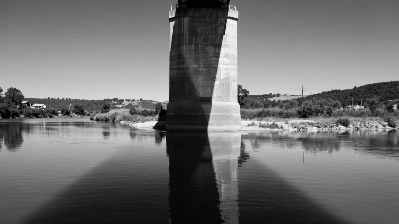 La telecamera si sposta verso l'alto, rivelando un pilone del ponte nel fiume per mostrare la sezione superiore e il riflesso della sua ombra.