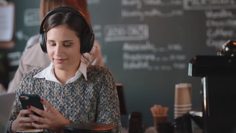 Mujer-Joven-Que-Usa-Un-Teléfono-Inteligente-En-Una-Cafetería-Enviando-Mensajes-De-Texto-Navegando-En-Línea-Disfrutando-Del-Entretenimiento-En-Las-Redes-Sociales-Escuchando-Música-En-Un-Restaurante-Concurrido