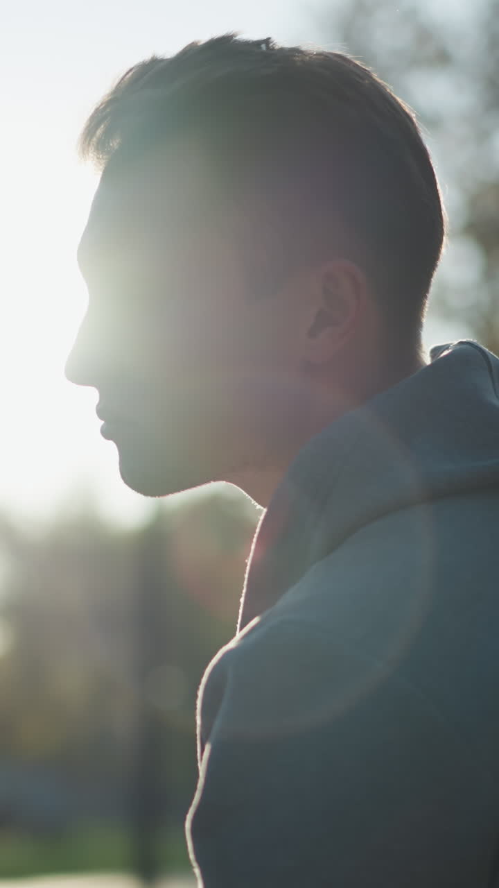 primer plano de un tipo con capucha gris, mirando hacia la derecha con una expresión pensativa parcialmente sombreada por la luz del sol, creando un estado de ánimo introspectivo. el fondo suavemente borroso con la luz del día brillante que destaca su cara