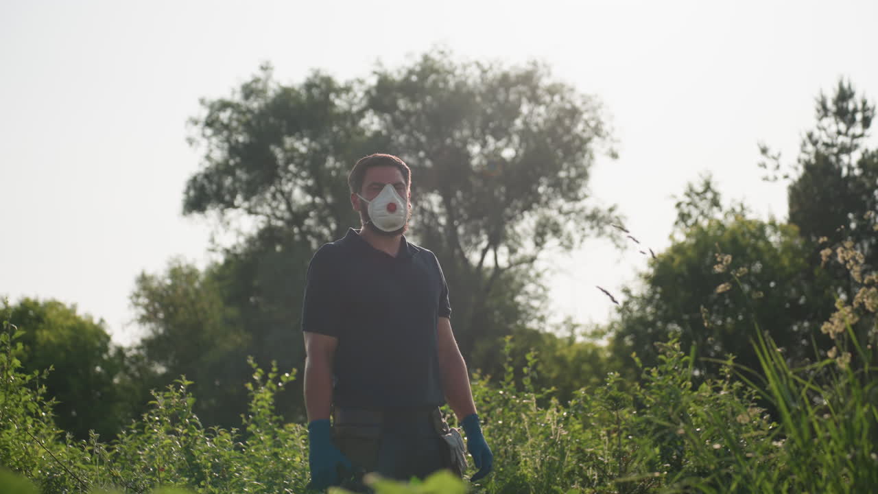 Gardener wearing protective mask and gloves stands amid green crops, gazing into distance under soft morning sunlight, portraying awareness of safety and responsibility in agricultural environment