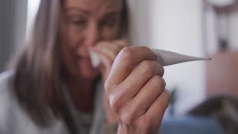 Woman-holding-a-thermometer-while-blowing-her-nose-at-home