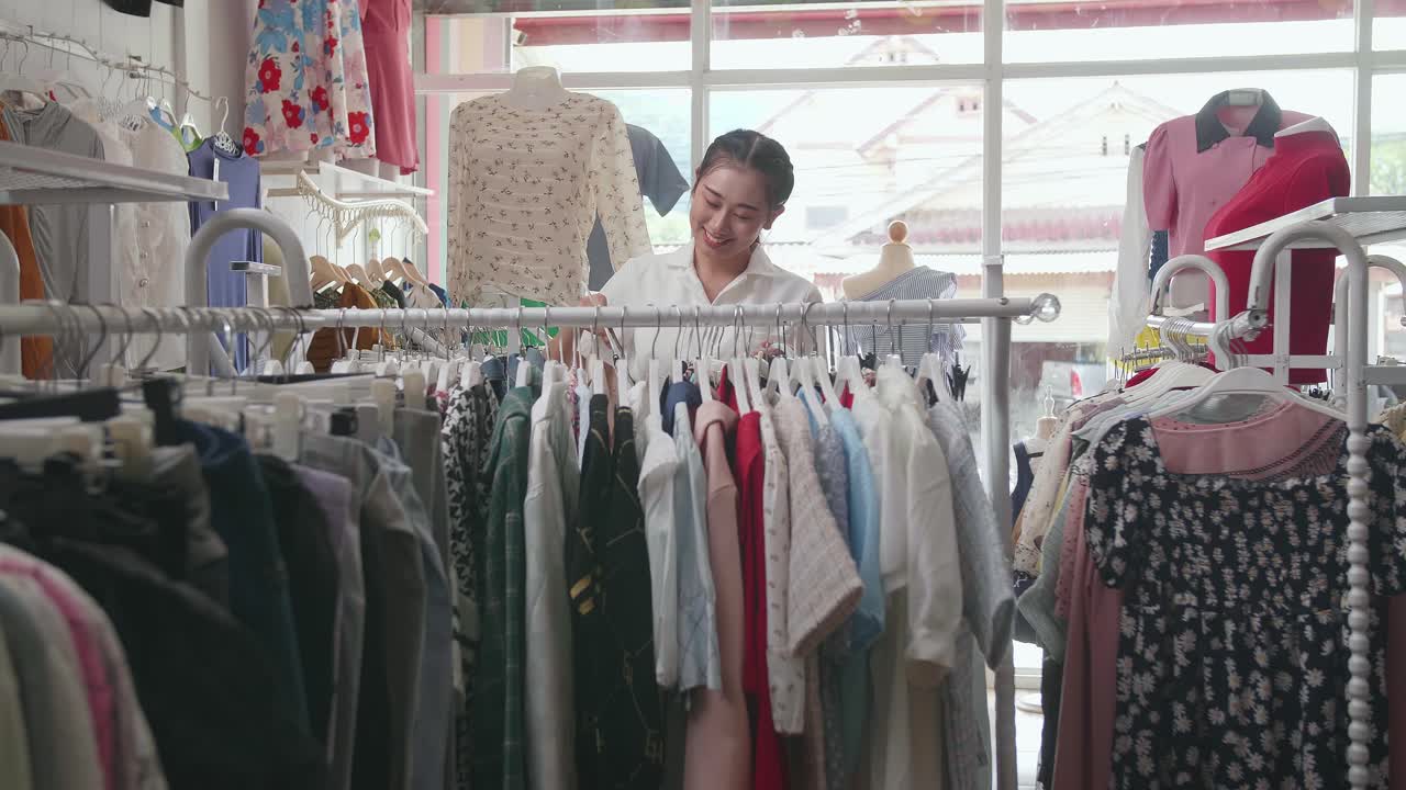 옷 가게 의 아시아인 여성 소유자 가 가게 에서 재고 를 확인 하는 동안 레일 에 드레스 를 매달고 있다