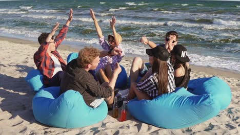 un grupo de jóvenes amigos sentados en la playa, divirtiéndose y dándose unos a otros los cinco en una noche de verano en cámara lenta