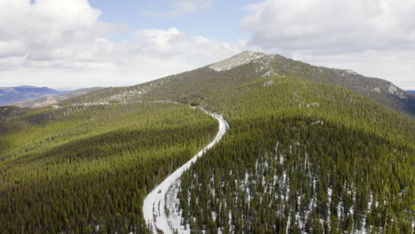 aerial reveal backward of sunny and snow covered back roads in the colorado mountains surrounded by bright green pine tree forests with blue skies