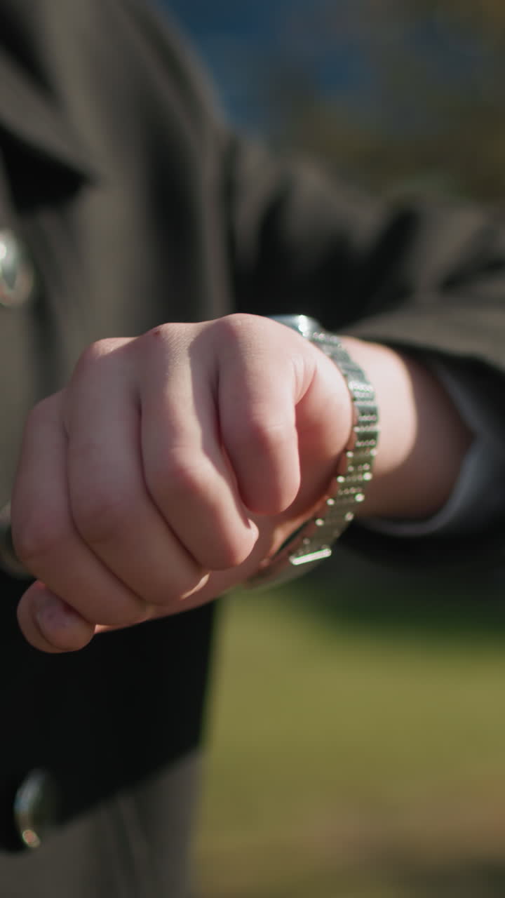 Close-up of elegant black coat featuring shiny metallic buttons in outdoor urban setting as individual checks wristwatch, background includes modern buildings, parked cars, and bright lighting