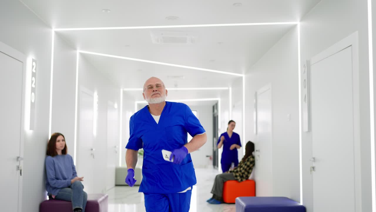 A hurrying older man doctor in a blue uniform runs along the bright corridor of the clinic holding a phone in his hands. An elderly man doctor with a beard runs along the corridor of a modern on-call clinic