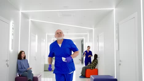 a hurrying older man doctor in a blue uniform runs along the bright corridor of the clinic holding a phone in his hands. an elderly man doctor with a beard runs along the corridor of a modern on-call clinic