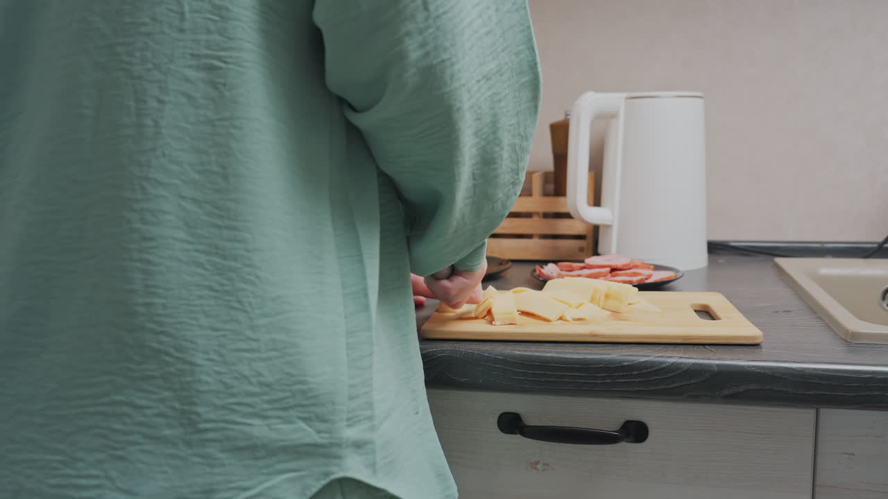 rear view of woman in green shirt slicing yellow cheese on cutting board in home kitchen with sliced meat on plate, electric kettle, spice jars in wooden holder near sink in soft daylight interior