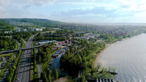 Antenne-über-Einer-Kleinen-Stadt-In-Einem-Europäischen-Land-Mit-Vielen-Lastwagen,-Die-Auf-Der-Schnellstraße-Fahren-Und-Waren-In-Der-Nähe-Eines-Großen-Sees-Liefern,-Seeschifffahrtspakete-Aus-Dem-Ausland,-Die-Neue-Standorte-Transportieren,-Grüne-Autos