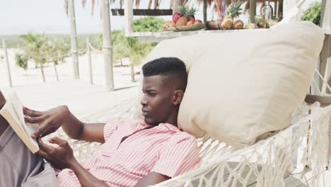 Feliz-Hombre-Afroamericano-Sentado-En-Una-Hamaca-Leyendo-Un-Libro-En-Una-Playa-Soleada,-Cámara-Lenta