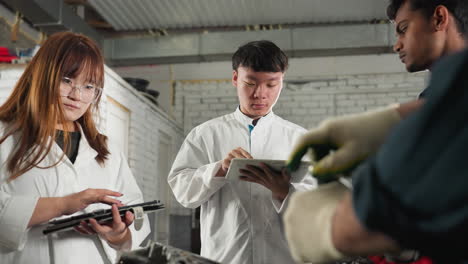 mechanic in white gloves demonstrates spanner use to students in lab coats, who take notes on tablets in automotive workshop, industrial workspace filled mechanical tools in background