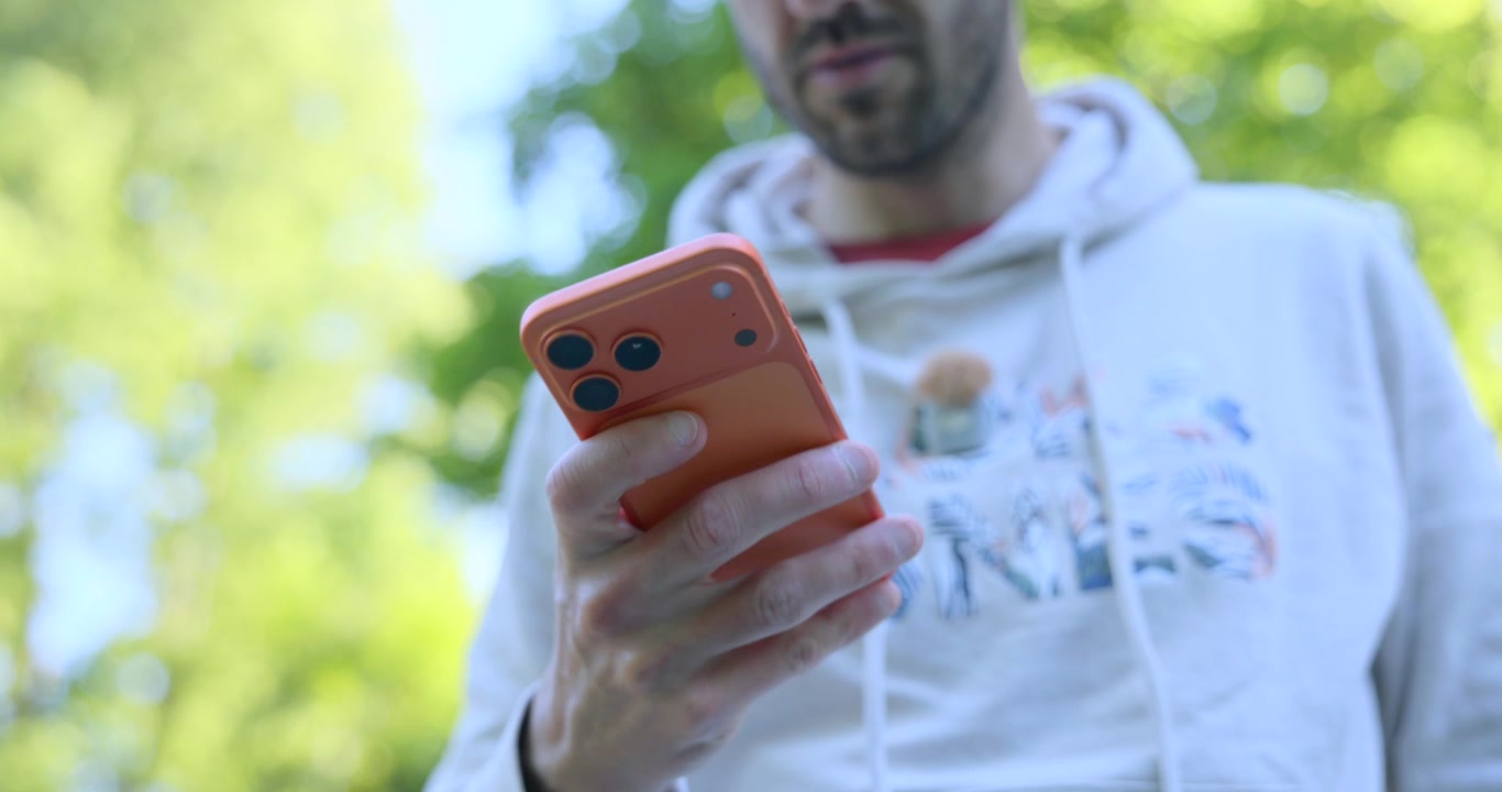 Low angle shot of a young man in a hoodie checking his mobile phone while walking in a park. He is scrolling through social media