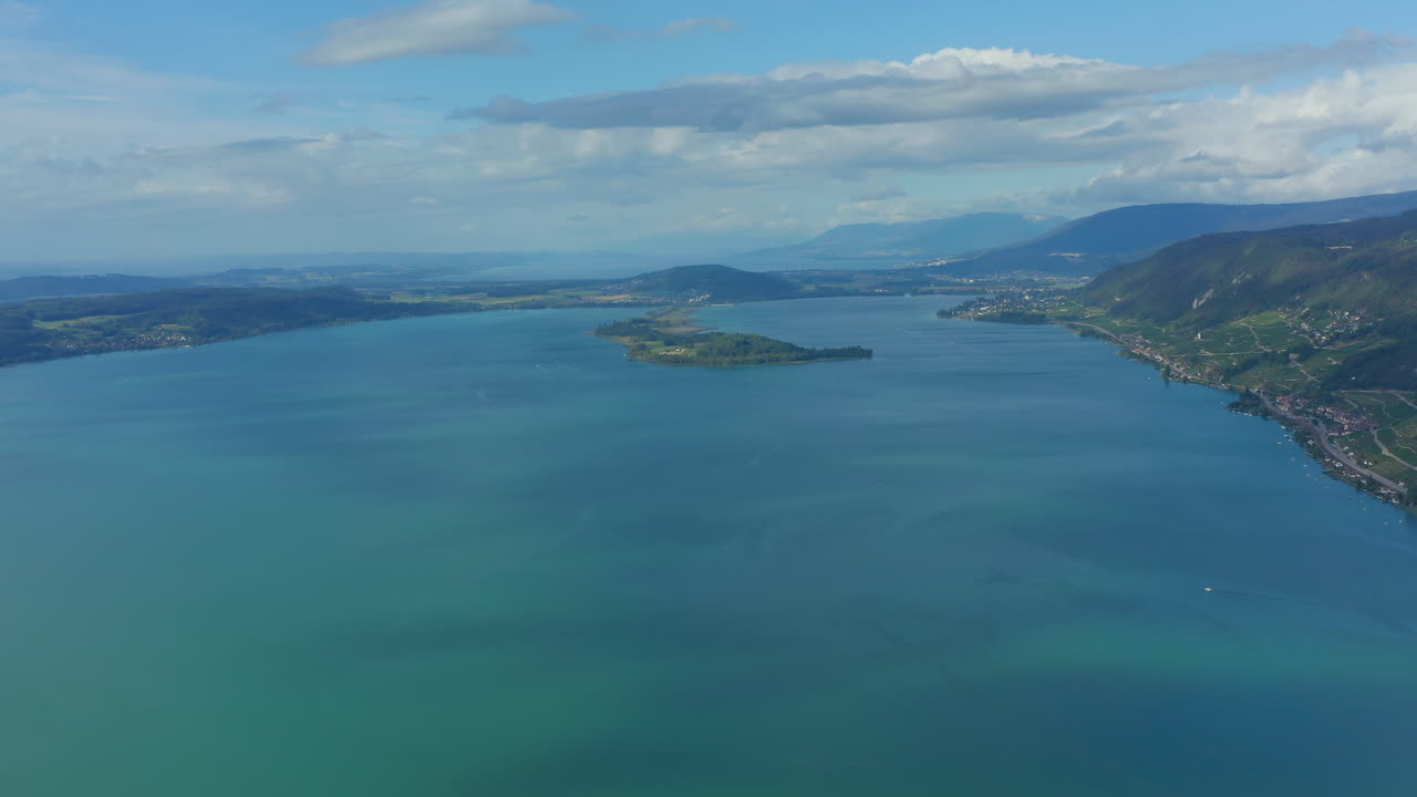 Aerial view of 'Lake Biel' in Switzerland in with clear greenish-blue water surrounded by a scenic landscape on a sunny day. 'Peterinsel' in the background under blue sky with fluffy clouds