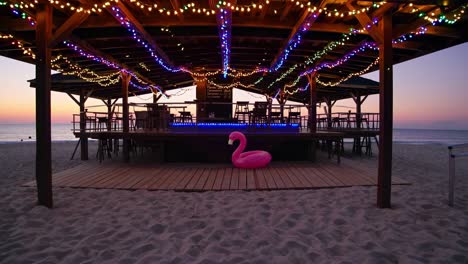 wooden beach bar with colorful string lights illuminating the counter and a pink flamingo inflatable at sunset, creating a serene and inviting atmosphere