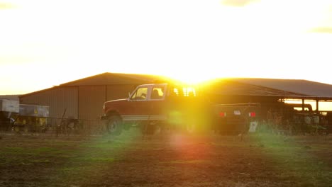 Drohnenaufnahme-Eines-Lastwagens-Und-Einer-Scheune-Inmitten-Eines-Orangefarbenen-Sonnenuntergangs,-Die-Drohne-Steigt-In-Die-Höhe-Und-Behält-Dabei-Den-Blick-Auf-Den-Lastwagen