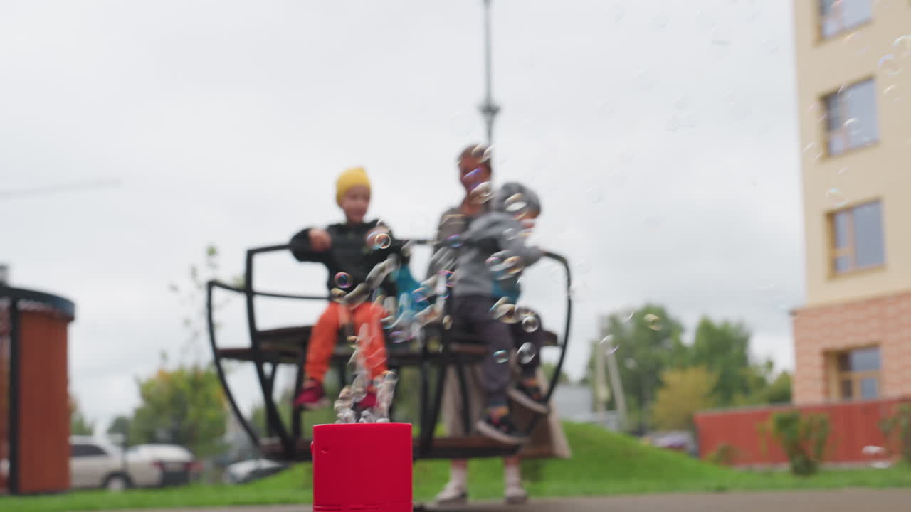 Blur background shows woman rotating children on merry go round anticlockwise while red bubble machine blows soap bubbles in foreground, cloudy sky over park, playful motion
