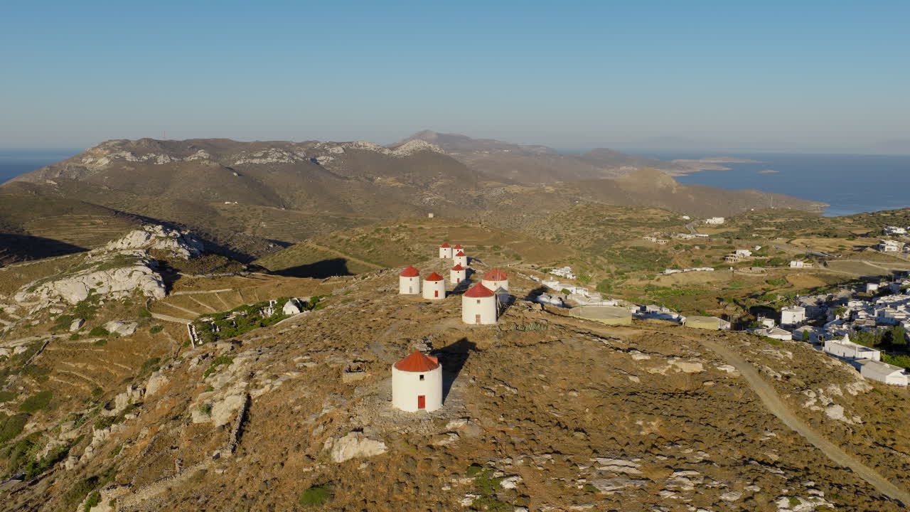 Drone footage flying over the traditional white windmills of Amorgos Chora at sunrise, bathed in soft golden morning light