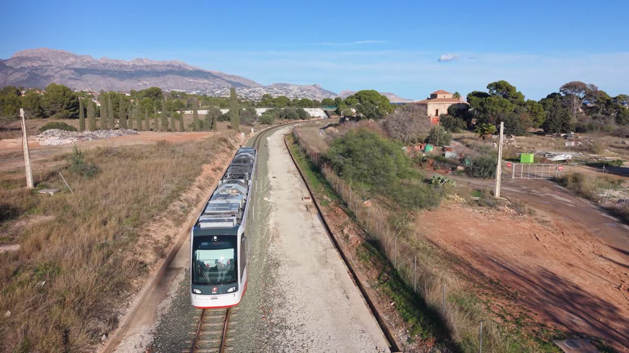 Albir, Costa Blanca, Spagna: treno che passa sulla linea da Benidorm ad Albir attraverso la campagna spagnola con montagne sullo sfondo, cielo azzurro limpido e sole splendente.