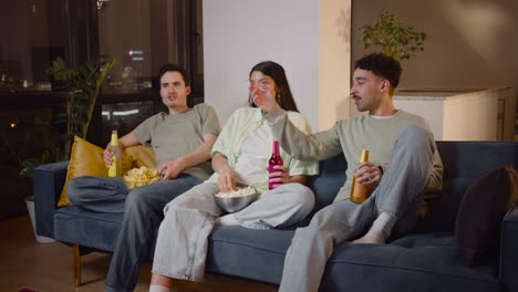 Tres-Amigos-Viendo-Una-Película-Interesante-En-La-Televisión-Sentados-En-El-Sofá,-Comiendo-Palomitas-De-Maíz-Y-Papas-Fritas-Y-Bebiendo-Refrescos