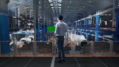 full body back view of an asian male professional worker standing at the center of the wind turbine factory, typing on laptop's keyboard with green screen and look at the factory once