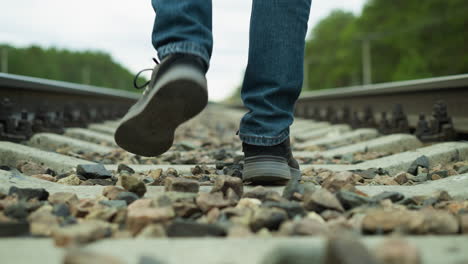 close leg view of a man in jeans and canvas shoes, walking calmly on rocky railway tracks, with a blur view of trees and electric poles