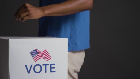 Close-Up-Of-Man-Casting-Vote-Into-American-Election-Ballot-Box-With-USA-Flag-Against-Black-Background-In-Slow-Motion