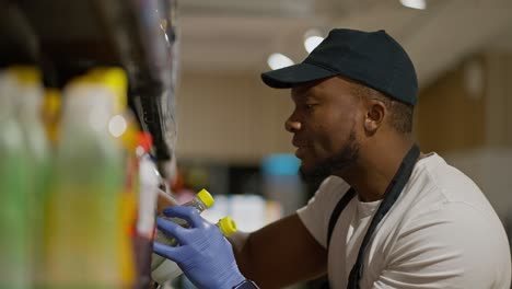 A-happy-man-with-Black-skin-in-a-black-cap-and-a-white-T-shirt-a-supermarket-employee-lays-out-and-sorts-bottles-in-a-special-display-case.-In-a-large-grocery-store