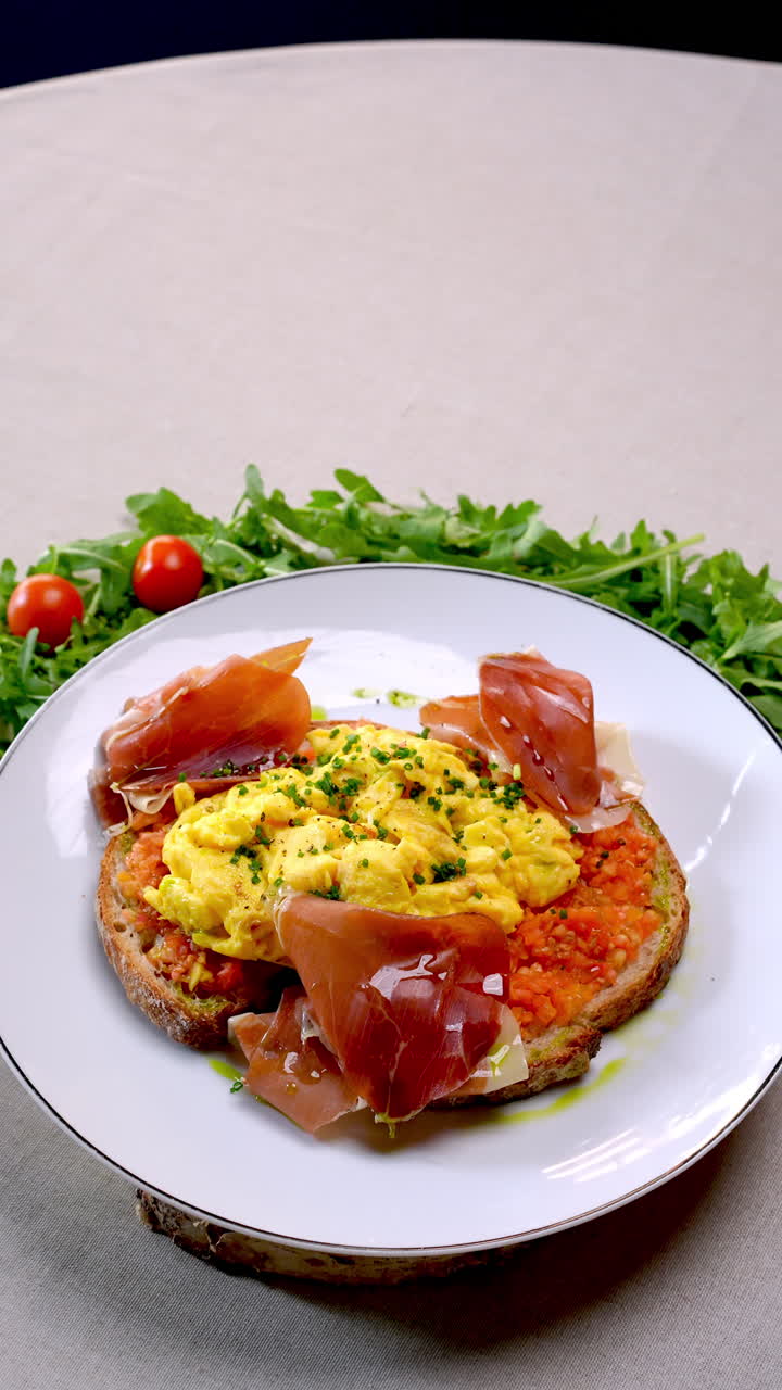 Scrambled eggs seasoned with chives lie on a bed of tomato sauce spread on toasted bread, accompanied by slices of prosciutto and garnished with arugula and cherry tomatoes