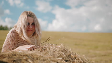 una mujer con gafas de sol se inclina para oler el heno bajo la cálida luz del sol en vastas tierras de cultivo, campo abierto, balas de heno esparcidas y cielo azul crean una atmósfera rural pacífica