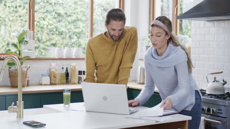 Diverse-couple-using-laptop-and-reading-documents-in-kitchen-at-home,-in-slow-motion