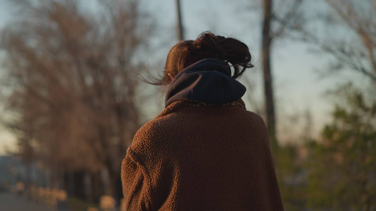 A somber girl walking through a park, with the wind gently blowing her hair. She is dressed in a brown coat and a hoodie, her expression reflecting deep thought