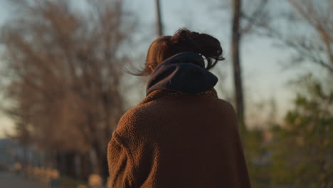 a somber girl walking through a park, with the wind gently blowing her hair. she is dressed in a brown coat and a hoodie, her expression reflecting deep thought