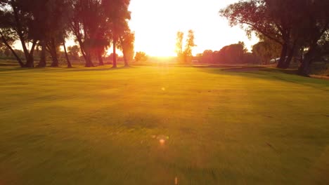 tiro de dron de ángulo bajo volando rápido hacia atrás sobre el campo de golf durante la puesta de sol entre árboles y estanque