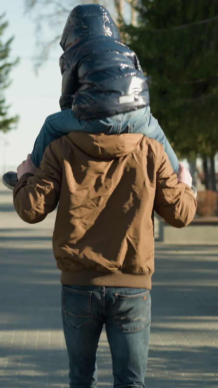 A father runs along a tree-lined paved path while carrying his child on his shoulders, the father holds the child's legs to maintain balance, both dressed in jackets