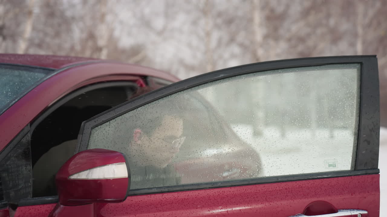Young man wearing winter jacket steps out of red car with visible water droplets on window and mirror during cold snowy weather with bare trees in background