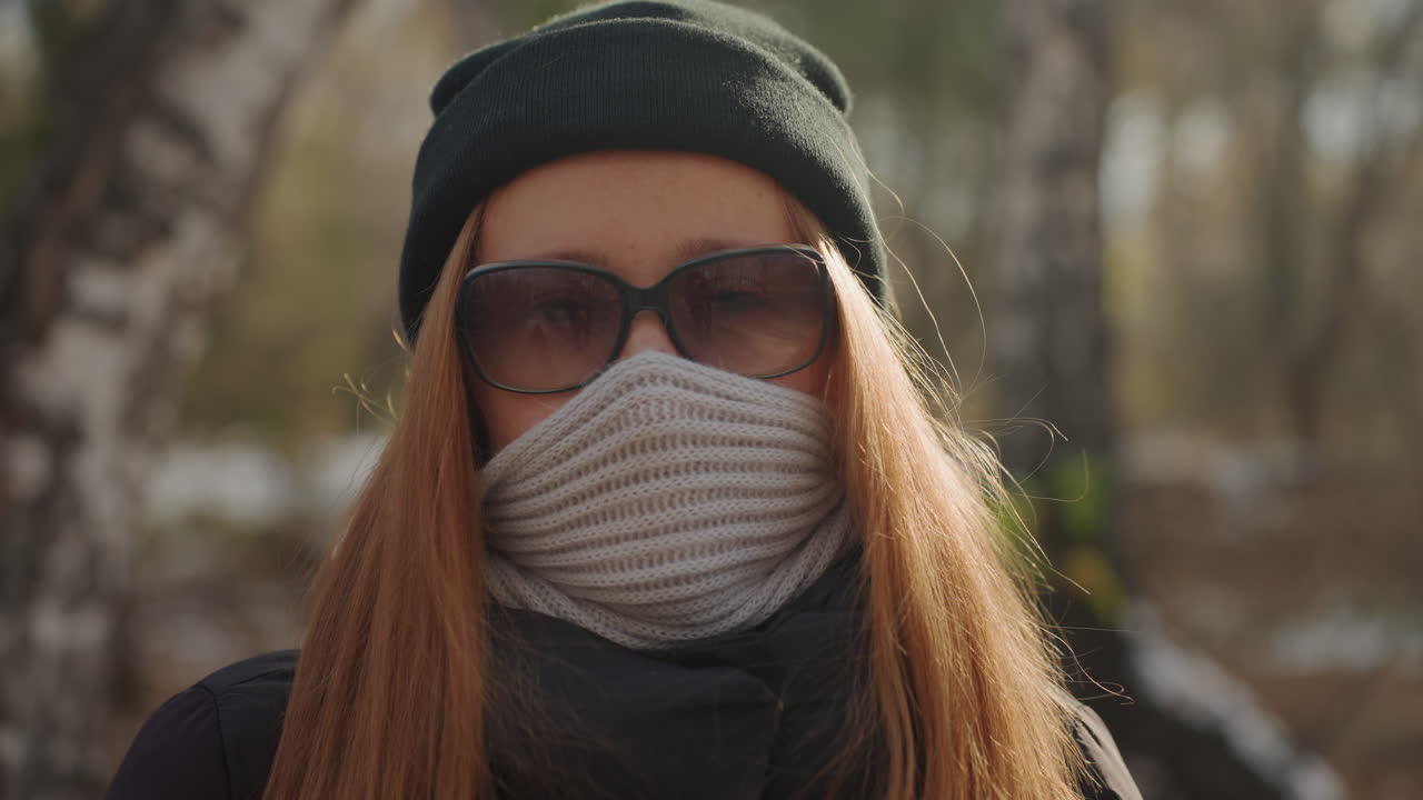 Mulher branca ruiva caminha por caminho de bétulas com máscara e gorro, closes de perfil e frontal mostram uma vibe de ladra ou andarilha pensativa, porém furtiva, folhas iluminadas pelo sol e tensão silenciosa em cenário natural
