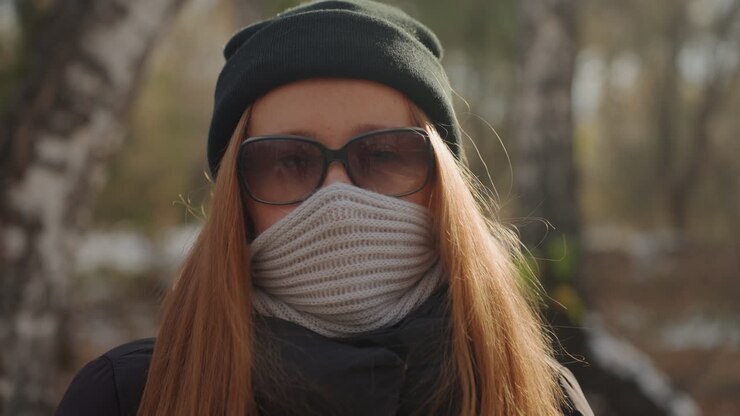 Redhaired white woman walks along birch path with mask and beanie, side profile and frontal closeups show reflective yet furtive thief or wanderer vibe, sunlit leaves and quiet tension in natural