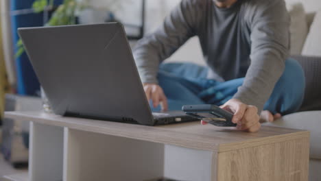 Young-adult-working-from-home-with-his-pajamas,-Uses-the-living-room-as-an-office-together-with-a-laptop,-He-is-distracted-by-a-reminder-popup-from-his-cell-phone-that-says-how-to-continue-today